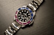 A luxury wristwatch with a metallic strap and a black dial. The bezel is adorned with alternating blue and pink gemstones, and the dial features white hour markers and a date window.