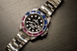 A luxury wristwatch with a metallic strap and a black dial. The bezel is adorned with alternating blue and pink gemstones, and the dial features white hour markers and a date window.