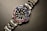 A luxury wristwatch with a metallic strap and a black dial. The bezel is adorned with alternating blue and pink gemstones, and the dial features white hour markers and a date window.