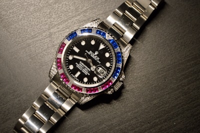 A luxury wristwatch with a metallic strap and a black dial. The bezel is adorned with alternating blue and pink gemstones, and the dial features white hour markers and a date window.