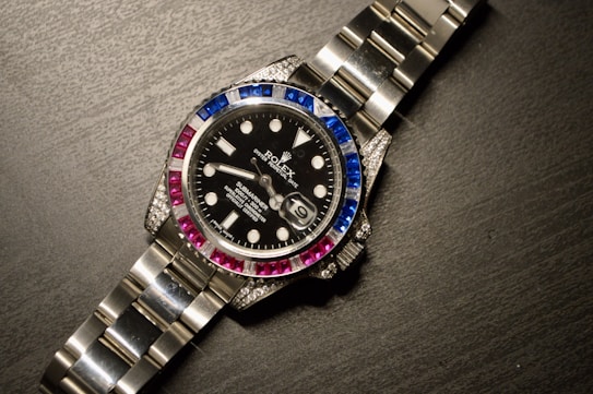 A luxury wristwatch with a metallic strap and a black dial. The bezel is adorned with alternating blue and pink gemstones, and the dial features white hour markers and a date window.