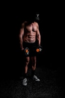 A muscular person stands shirtless in a dark environment, holding dumbbells with a focused posture. The individual wears a cap with a distinctive logo, shorts, and sneakers, suggesting a fitness or workout setting.