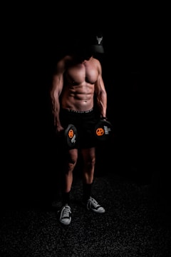 A muscular person stands shirtless in a dark environment, holding dumbbells with a focused posture. The individual wears a cap with a distinctive logo, shorts, and sneakers, suggesting a fitness or workout setting.