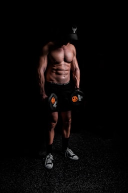 A muscular person stands shirtless in a dark environment, holding dumbbells with a focused posture. The individual wears a cap with a distinctive logo, shorts, and sneakers, suggesting a fitness or workout setting.
