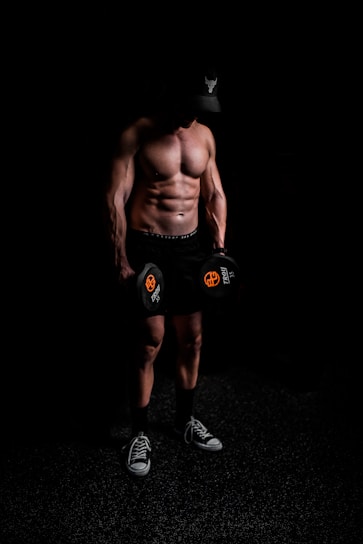 A muscular person stands shirtless in a dark environment, holding dumbbells with a focused posture. The individual wears a cap with a distinctive logo, shorts, and sneakers, suggesting a fitness or workout setting.
