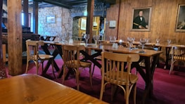 A cozy, rustic dining room features several wooden tables and chairs arranged neatly on a red carpet. The room is adorned with stone walls and wooden panels, giving it a warm, inviting ambiance. Wine glasses and table settings are in place, ready for guests. Two framed portraits hang on the wall, adding a classic touch to the decor.
