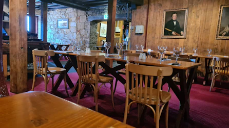 A cozy, rustic dining room features several wooden tables and chairs arranged neatly on a red carpet. The room is adorned with stone walls and wooden panels, giving it a warm, inviting ambiance. Wine glasses and table settings are in place, ready for guests. Two framed portraits hang on the wall, adding a classic touch to the decor.