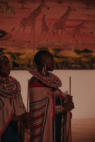 Two individuals in traditional attire featuring intricate beadwork and patterned fabrics stand in front of a mural depicting giraffes in a savannah landscape. The attire is adorned with colorful beaded necklaces and wraps.
