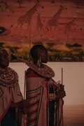 Two individuals in traditional attire featuring intricate beadwork and patterned fabrics stand in front of a mural depicting giraffes in a savannah landscape. The attire is adorned with colorful beaded necklaces and wraps.