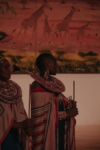 Two individuals in traditional attire featuring intricate beadwork and patterned fabrics stand in front of a mural depicting giraffes in a savannah landscape. The attire is adorned with colorful beaded necklaces and wraps.