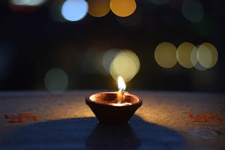A small clay diya lamp is lit with a flickering flame, casting a warm glow on the surface it rests upon. The background features soft, colorful bokeh lights in shades of blue and yellow, creating a serene atmosphere.