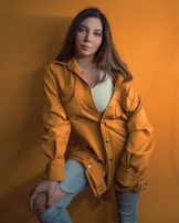 A person wearing a mustard yellow jacket and a white top poses against a matching mustard yellow backdrop. They have long, dark hair and light makeup. Their jeans are light blue and slightly ripped at the knee.
