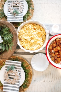 A rustic table setting with a central focus on a dish of macaroni and cheese in a white ceramic bowl, next to a red-and-white bowl of fried chicken pieces. The table is decorated with woven placemats, striped napkins, and greenery with pine cones, topped with snowflake decorations. There are also white plates arranged around the table setting.
