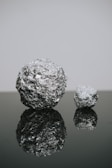 Two crumpled balls of aluminum foil are placed on a reflective surface, one larger and one smaller. The texture of the foil creates a shiny and detailed pattern. The reflection on the dark surface adds depth to the composition.