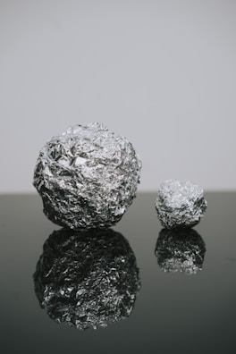Two crumpled balls of aluminum foil are placed on a reflective surface, one larger and one smaller. The texture of the foil creates a shiny and detailed pattern. The reflection on the dark surface adds depth to the composition.