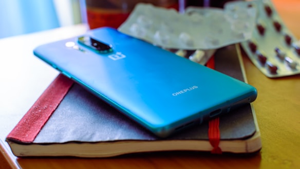A blue smartphone placed on top of a closed red and black notebook. In the background, a blister pack of tablets and a cup are partially visible, suggesting a casual or workspace environment.