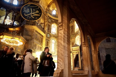 A large, ornate interior space featuring intricate Islamic calligraphy and architectural elements such as arched windows and a chandelier. Several people are present, including a man taking photos and others admiring the surroundings. The atmosphere is warm, with golden tones illuminating the stone surfaces.
