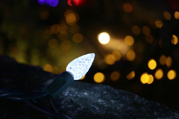 A close-up view of a single white LED bulb on a string of lights, set against a dark background with softly blurred, out-of-focus lights in the distance creating a bokeh effect.
