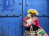 A young person in traditional colorful attire is holding a small white lamb. The person is wearing a vibrant pink top and a skirt with intricate multicolored patterns. A yellow woven hat with fringe adorns their head, and a blue wooden door serves as a backdrop.