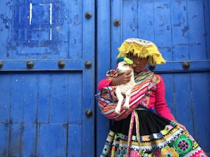 A young person in traditional colorful attire is holding a small white lamb. The person is wearing a vibrant pink top and a skirt with intricate multicolored patterns. A yellow woven hat with fringe adorns their head, and a blue wooden door serves as a backdrop.