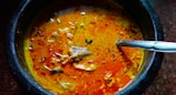 A vibrant curry is served in a dark stone or ceramic bowl. The dish features a rich, orange-red broth with visible chunks of meat and garnished with herbs and spices. A stainless steel spoon rests inside the bowl, ready for use.