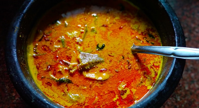 A vibrant curry is served in a dark stone or ceramic bowl. The dish features a rich, orange-red broth with visible chunks of meat and garnished with herbs and spices. A stainless steel spoon rests inside the bowl, ready for use.