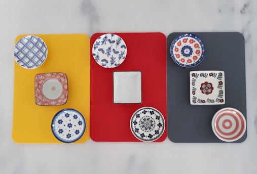 A display of nine colorful, patterned ceramic plates and dishes arranged on three rectangular mats in yellow, red, and dark gray. Each mat has three dishes with various designs, including floral, geometric, and abstract patterns in colors like blue, red, black, and white.