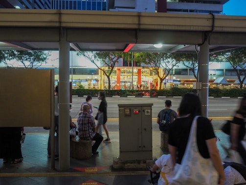 A bus stop scene in an urban area with several people sitting and standing, some using mobile devices. The background features a busy street lined with trees and a brightly lit shopping mall. Signs and lights create a modern city atmosphere.