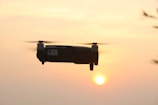 A drone flies against the backdrop of a setting sun, with softly blurred rotor blades in motion. The sky is a gradient of warm colors from orange to pale yellow, indicating a calm evening scene. The drone is centrally positioned, casting a silhouette framed against the light of the sun.