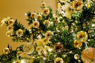 A festive Christmas tree adorned with a variety of golden and metallic ornaments, including shiny spheres and intricate ribbon bows. The evergreen branches are illuminated by soft, warm lights, enhancing the luxurious and festive appearance.