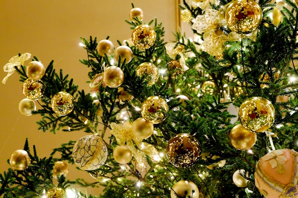 A festive Christmas tree adorned with a variety of golden and metallic ornaments, including shiny spheres and intricate ribbon bows. The evergreen branches are illuminated by soft, warm lights, enhancing the luxurious and festive appearance.