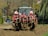 A large tractor with a red plow attachment is actively tilling a field. The equipment is covered in freshly turned earth, indicating recent use. The background features a row of trees and hedges along a residential street, with a parked car visible. The scene depicts a rural area transitioning into an urban setting.