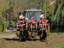 A large tractor with a red plow attachment is actively tilling a field. The equipment is covered in freshly turned earth, indicating recent use. The background features a row of trees and hedges along a residential street, with a parked car visible. The scene depicts a rural area transitioning into an urban setting.