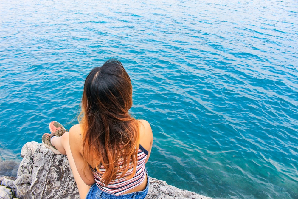 woman in white and blue stripe tank top sitting on rock near body of water during