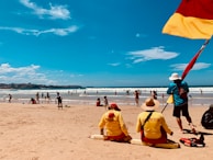 Lifeguards wearing yellow uniforms sit on the sandy beach, monitoring swimmers in the ocean. A flagpole with a red and yellow flag stands next to one of the lifeguards. Several people are scattered across the beach and in the water, enjoying a sunny day. The sky is clear with a few clouds, and the sea is calm with gentle waves.