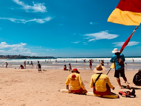 Lifeguards wearing yellow uniforms sit on the sandy beach, monitoring swimmers in the ocean. A flagpole with a red and yellow flag stands next to one of the lifeguards. Several people are scattered across the beach and in the water, enjoying a sunny day. The sky is clear with a few clouds, and the sea is calm with gentle waves.