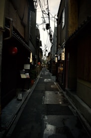 A narrow alleyway lined with traditional buildings, with numerous overhead power lines. The street is wet, reflecting light from various signs and lamps, creating a moody atmosphere.
