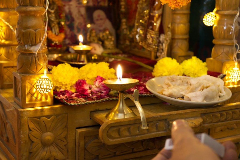 A traditional altar adorned with vibrant marigold and rose flowers, featuring small oil lamps with flickering flames. The setup includes a brass plate containing white ceremonial offerings, placed on an intricately carved wooden surface. Soft, warm lighting adds to the sacred atmosphere.