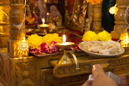 A traditional altar adorned with vibrant marigold and rose flowers, featuring small oil lamps with flickering flames. The setup includes a brass plate containing white ceremonial offerings, placed on an intricately carved wooden surface. Soft, warm lighting adds to the sacred atmosphere.