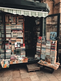 A small bookshop stall displaying a variety of books stacked in piles both inside and outside. The shop has an awning, and the books are arranged neatly across shelves and wooden pallets on the ground. Some book covers are partially visible, showing a mix of colors and book genres.