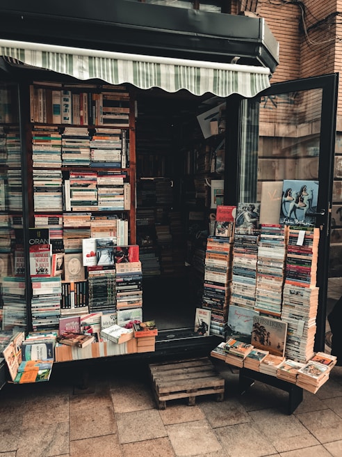 A small bookshop stall displaying a variety of books stacked in piles both inside and outside. The shop has an awning, and the books are arranged neatly across shelves and wooden pallets on the ground. Some book covers are partially visible, showing a mix of colors and book genres.