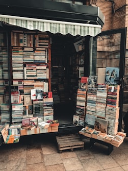 A small bookshop stall displaying a variety of books stacked in piles both inside and outside. The shop has an awning, and the books are arranged neatly across shelves and wooden pallets on the ground. Some book covers are partially visible, showing a mix of colors and book genres.