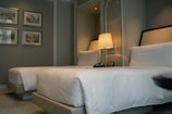 A well-designed hotel room featuring two neatly made twin beds with white linens. A nightstand sits between the beds, holding a lamp, telephone, and a clock. Four framed artworks hang on the wall, adding a touch of elegance to the space. The room exudes a calm and sophisticated ambiance with its neutral color palette.