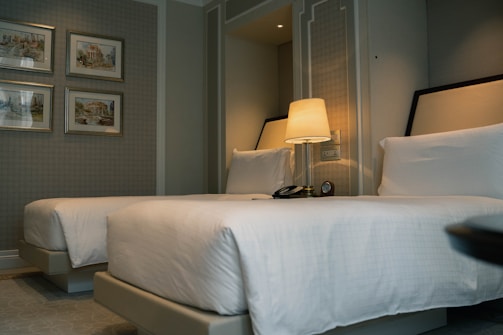 A well-designed hotel room featuring two neatly made twin beds with white linens. A nightstand sits between the beds, holding a lamp, telephone, and a clock. Four framed artworks hang on the wall, adding a touch of elegance to the space. The room exudes a calm and sophisticated ambiance with its neutral color palette.