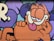 A colorful mural featuring a cartoon cat with orange fur and black stripes. The cat is depicted with a relaxed expression, its eyes half-closed, and its tongue playfully sticking out. The background incorporates purple and blue hues with bold outlines.