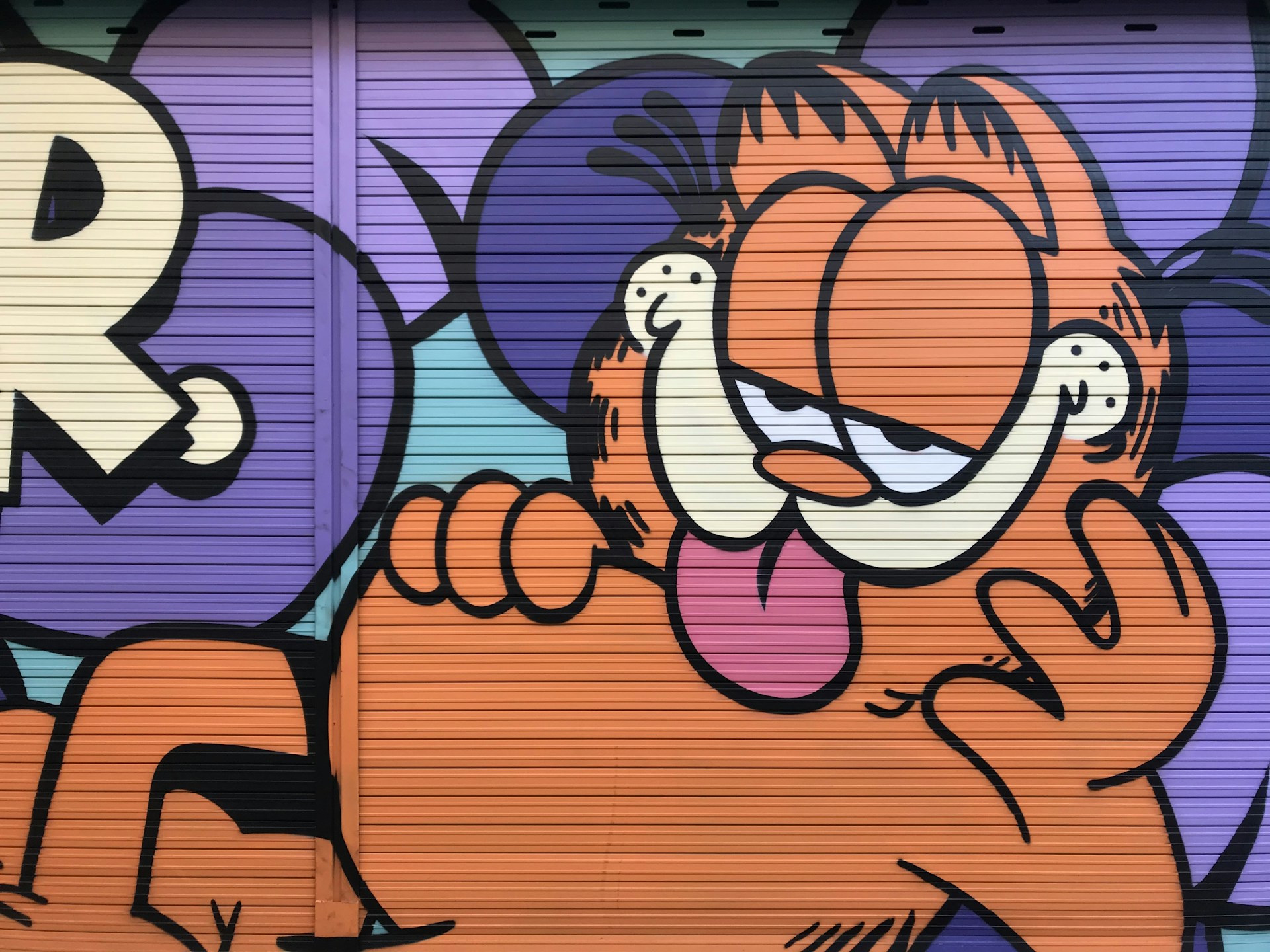A colorful mural featuring a cartoon cat with orange fur and black stripes. The cat is depicted with a relaxed expression, its eyes half-closed, and its tongue playfully sticking out. The background incorporates purple and blue hues with bold outlines.