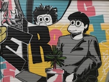 A vibrant mural featuring two cartoon-like characters with exaggerated eyes, one with spiky hair and glasses, and the other with a smooth hairstyle. They stand in front of large colorful letters on a bright background, with a small plant in a pot in front of them.