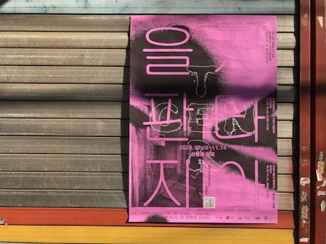 A pink poster with Korean text is affixed to a corrugated metal shutter. Black and white graphics and text accompany the vibrant pink color. The poster includes a date range and QR code, suggesting an event or exhibition. The background is an industrial setting with subtle shadows cast over the metal surface.