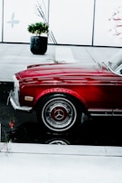 A polished, red vintage car with a chrome grille and mirror is prominently displayed indoors. The car is parked next to a black reflective surface, which creates a clear reflection of the car's wheel and bottom. In the background, there is a potted plant and a decorative wall with minimalist designs.