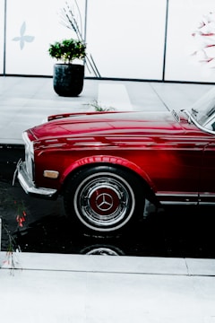 A polished, red vintage car with a chrome grille and mirror is prominently displayed indoors. The car is parked next to a black reflective surface, which creates a clear reflection of the car's wheel and bottom. In the background, there is a potted plant and a decorative wall with minimalist designs.